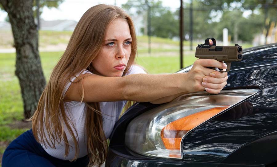 In this jpeg image, a young woman aims a Springfield Armory Hellcat pistol over the hood of a Toyota Camry while training. Her hands and fingers are wrapped around the grip while her hair is pulled back. She had blue eyes and is wearing a white shirt.